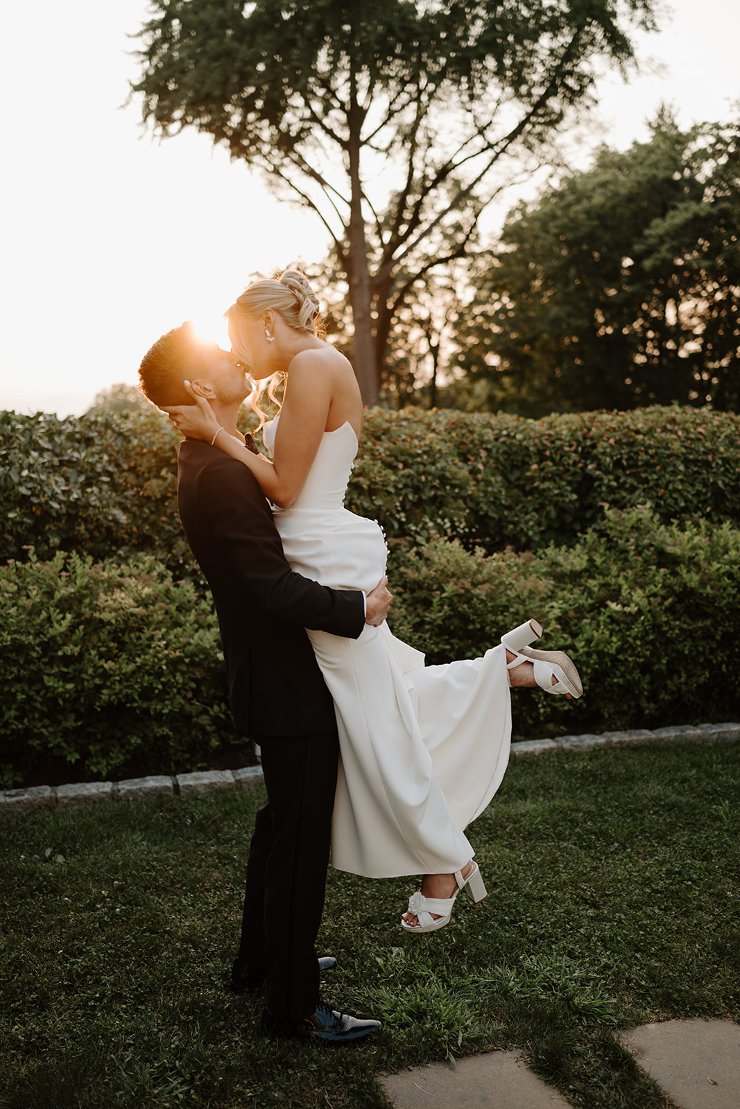 Groom lifts and kisses the bride in a garden at sunset, with warm golden light illuminating the moment.